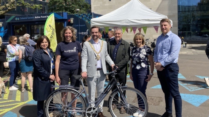 A group of men and women stand outside in the sun with the Lord Mayor of Belfast in the middle, wearing his gold chain of office and holding a bicycle