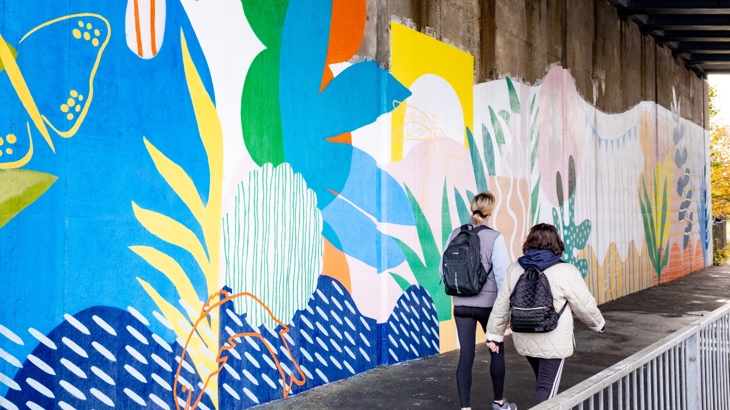 Brightly coloured mural in underpass in Irvine with two people walking past looking up. Mural depicts local wildlife found in River Irvine.