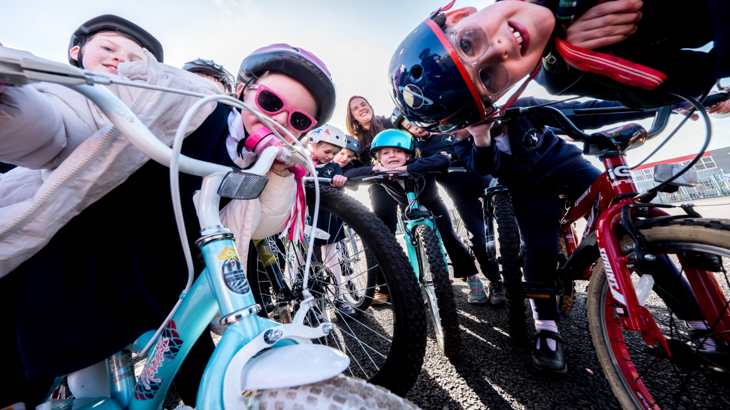 A group of Primary school children with cycles, helmets and sunglasses gathered around a camera on a playground smiling