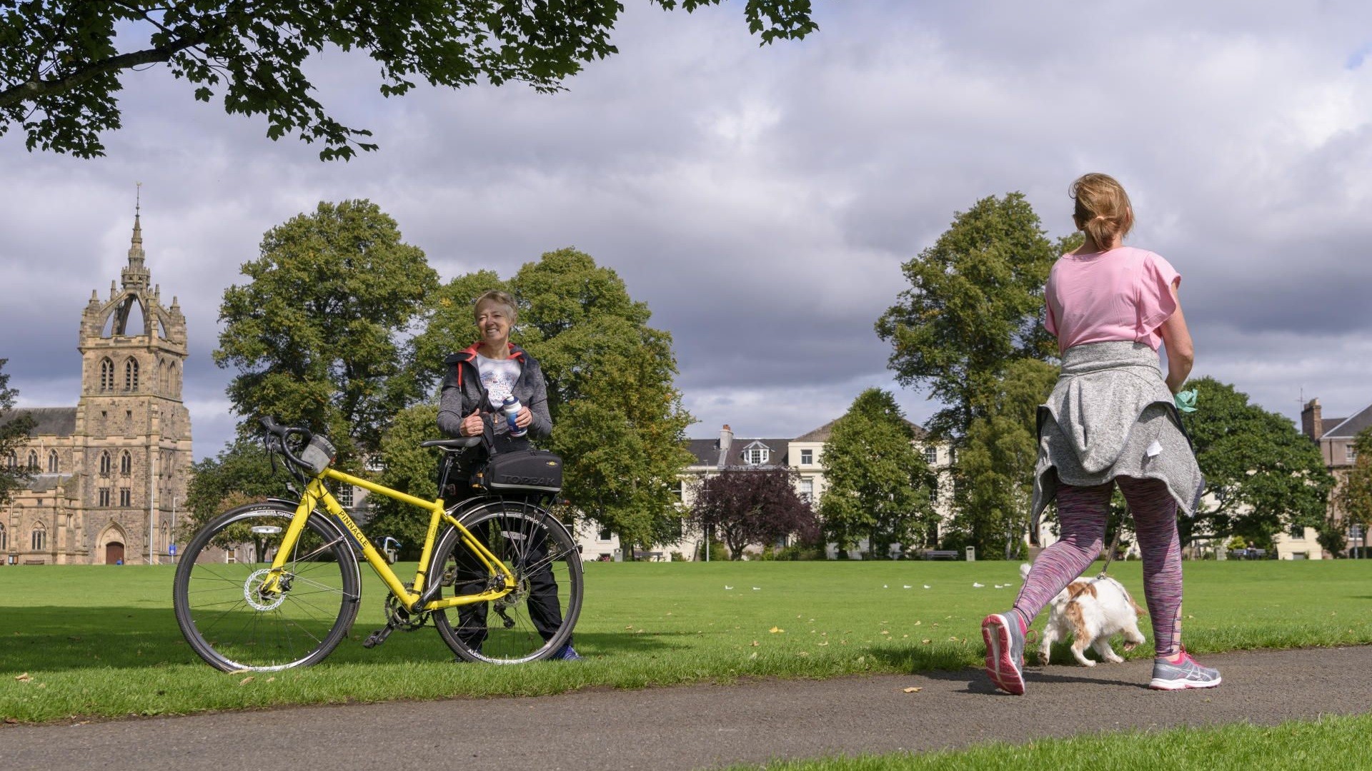 Woman cyclist on grass taking a drink as woman dog walker passes by
