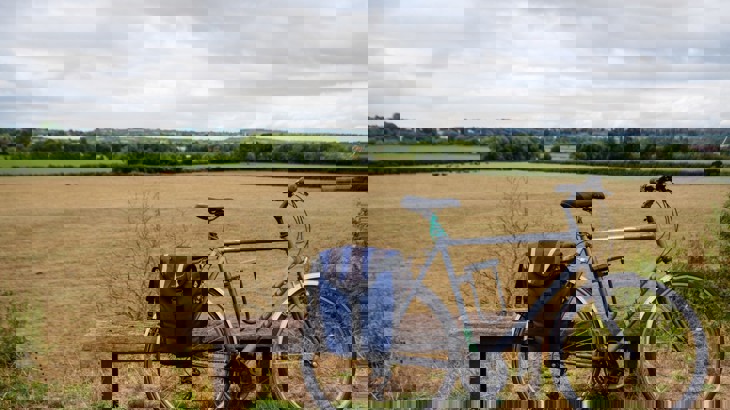 A road bike with a blue saddle bag is leant up against a backless wooden bench. Behind is a flat agricultural field on a sunny day in the countryside.