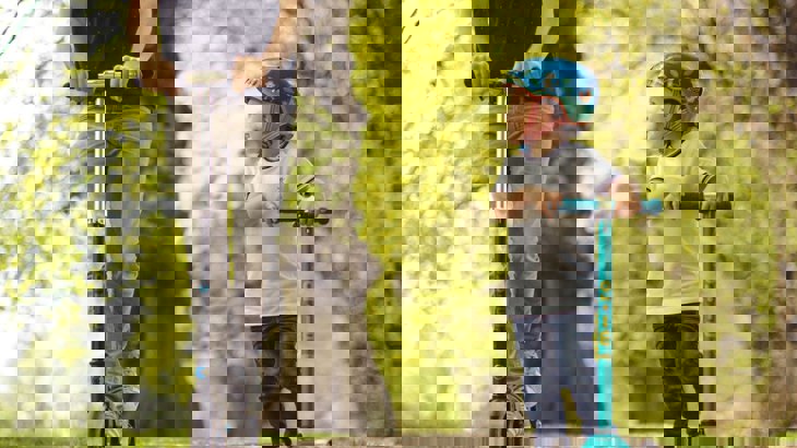 Boy in t shirt, jeans and helmet riding blue scooter with man riding black scooter through park