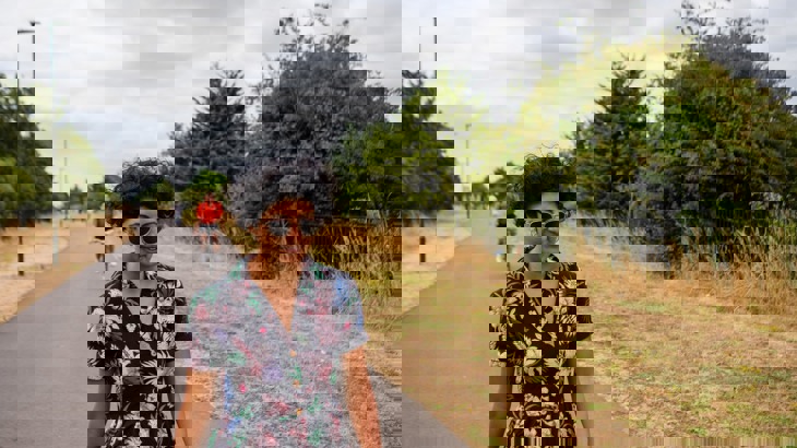 Woman in sunglasses walking on asphalt walking and cycling path with brown grass and green trees to the side and cyclist behind