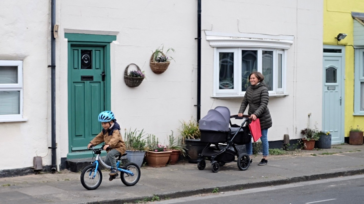 A woman pushing a pram along a bright residential road while her young son pedals ahead on a blue bike wearing a blue helmet