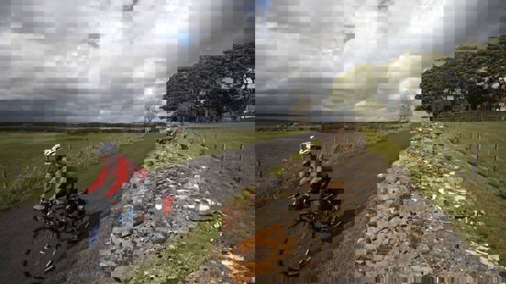 Cyclist on road bike with panniers wearing red jacket and white helmet cycling on road past Hadrian's Wall