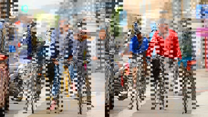Three adults wearing helmets and riding bikes are waiting at a traffic light in a cycle lane in a city, there are queuing cars beside them and everyone is smiling.