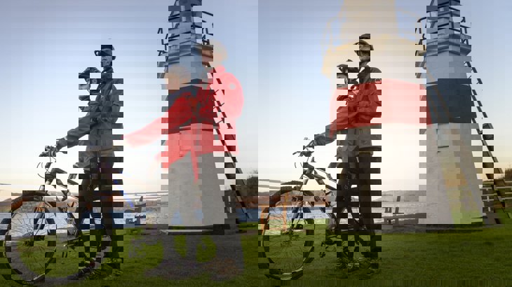 Two people in red coats and cycling helmets, one with mountain bike, standing in front of white and red lighthouse