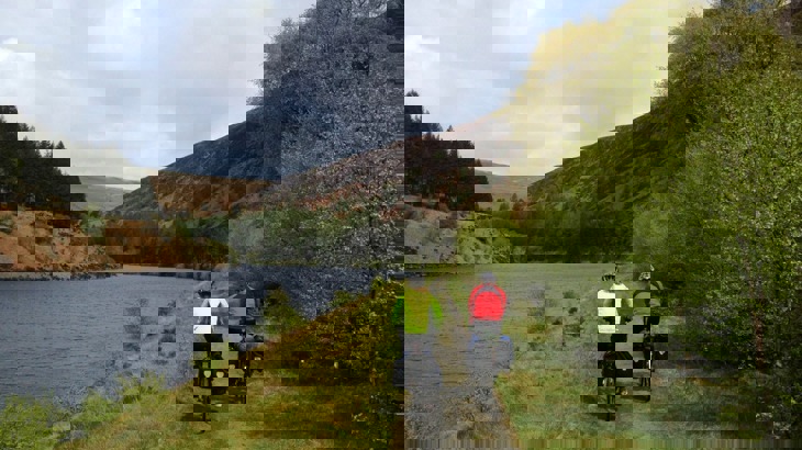 Two cyclists with touring gear on cycle path by river in valley with trees