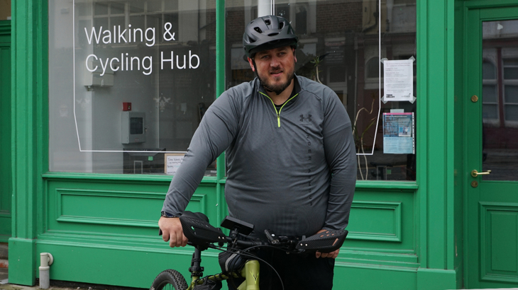 A man stood with his cycle wearing a helmet and dark clothing outside one of Walk Wheel Cycle Trust's walking and cycling hubs