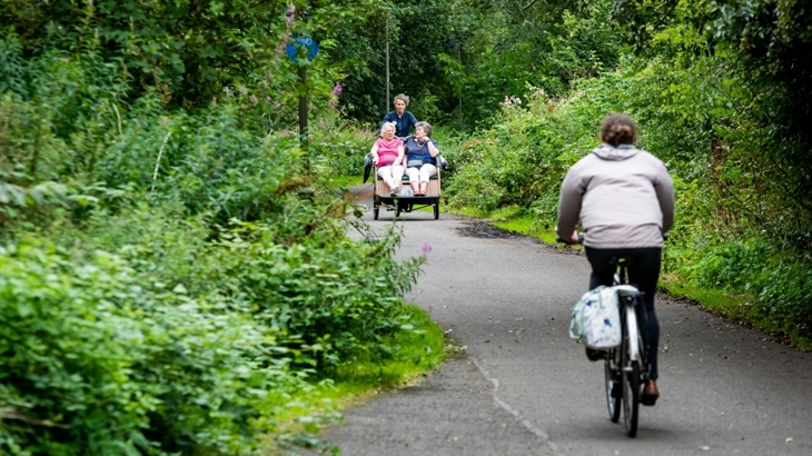 Two women are travelling on the front of a rickshaw along the Roseburn path, Edinburgh, a traffic free path of the National Cycle Network. A person on a bike is approaching them in the foreground. The scene is bright green with tall tress and bushes in full leaf.