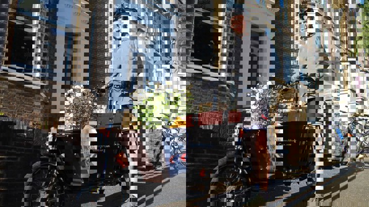 A man stands astride a cargo bike loaded with shopping. He is outdoors on a London street. He is smiling, the weather is fine and the mood is pleasant.