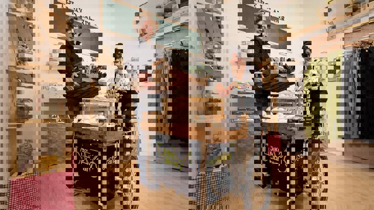 Man and woman standing inside a shop smiling next to a black, Walk Wheel Cycle Trust branded cargo bike they use for deliveries.