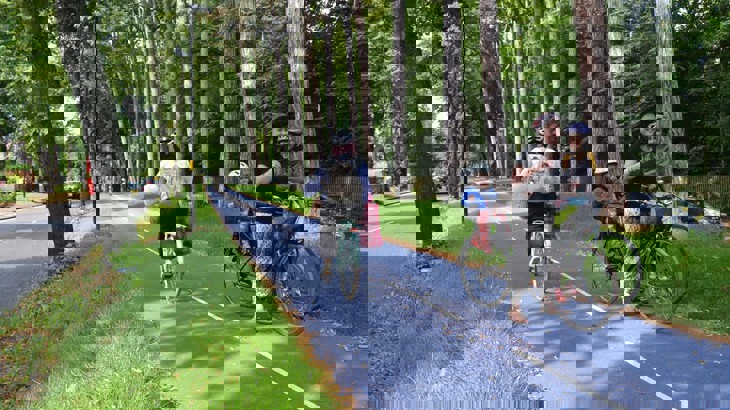 Person cycling a long a blue segregated cycle route lined with green trees as a family with bikes stop and chat to a friend next to the route.