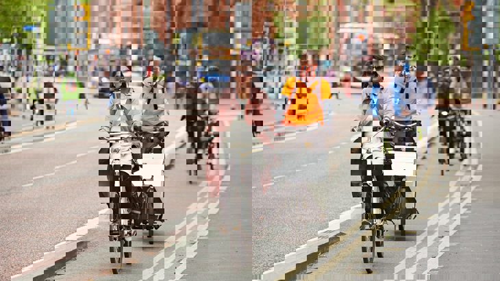 Woman wearing a pink jacket and sunglasses cycling down a segregated cycle lane in Manchester city centre with a man on a cargo bike behind her.
