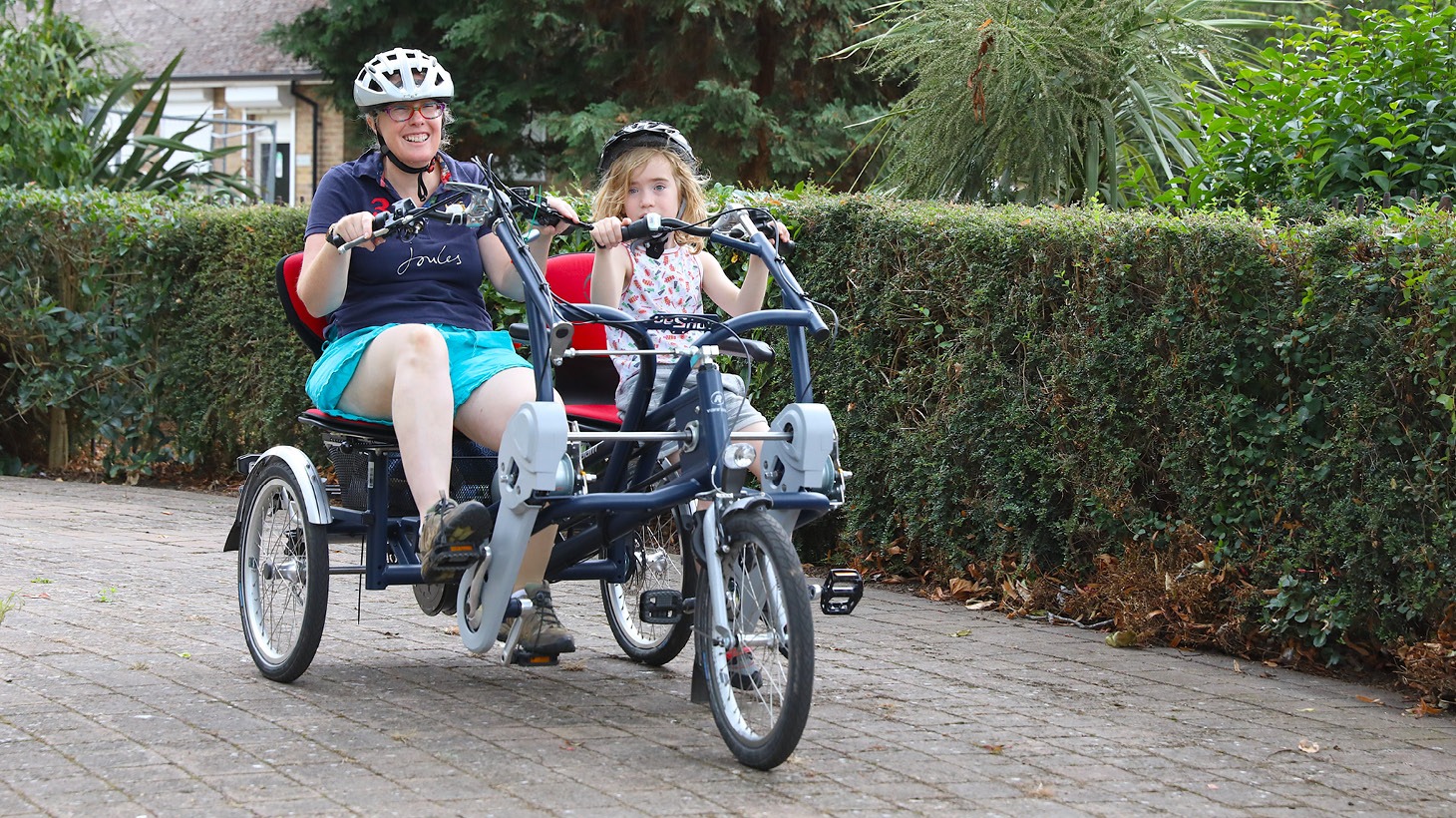 Mum and daughter wearing helmets, smiling as they ride an adapted two-person tricycle through a green park.
