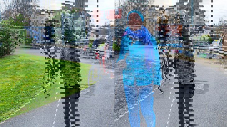 Lady in bright blue using a cane walking along a traffic-free path with a male cycling behind her.