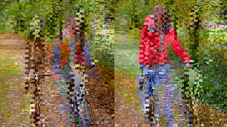 Two women in waterproof jackets and hats cycling through a park surrounded by crisp, orange and brown autumn leaves.
