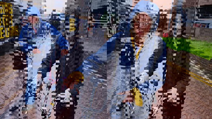 Lady wearing a navy blue waterproof smiling as she walks down a street with her bike in the rain.