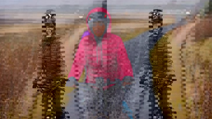 Lady wearing a red coat with the hood pulled up over her cycle helmet, cycling down a traffic-free path with grey skies in the background.