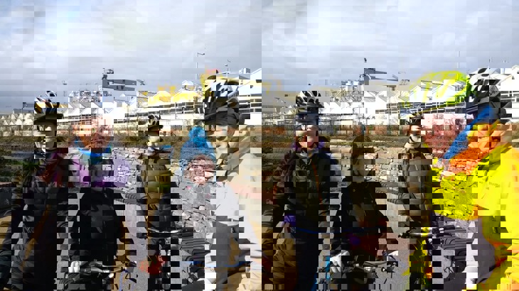 three women on a group ride with Walk Wheel Cycle Trust staff member Tom in a park with Belfast Harland and Wolf cranes in the background