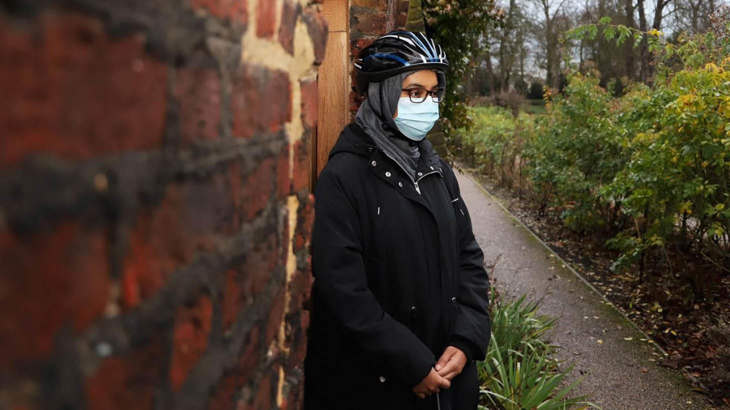 Shaima standing next to a brick wall in a local park, wearing a protective mask and a cycle helmet.