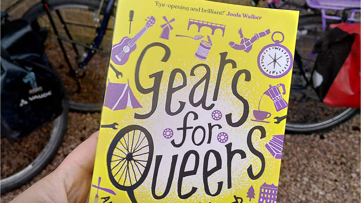 Close up of the front cover of Abi and Lilith's book, Gears for Queers, with a bicycle in the background.
