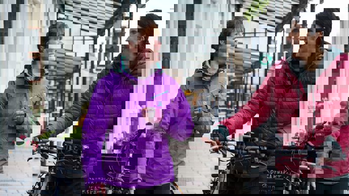 Two women wearing brightly coloured raincoats, chatting and walking with their bikes through a high street