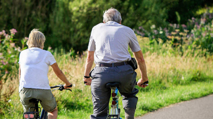 Two older people cycling away from the camera down a traffic-free path, surrounded by green grass and trees.