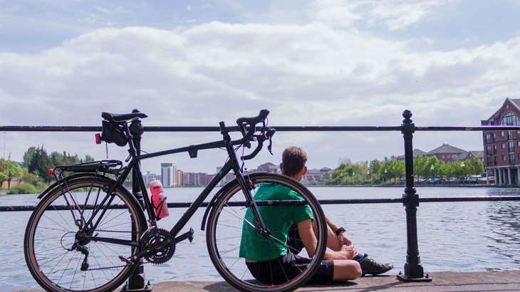 Man sitting alone with his bicycle propped up on the railings next to him, overlooking a beautiful sunset on a canal in Cardiff.