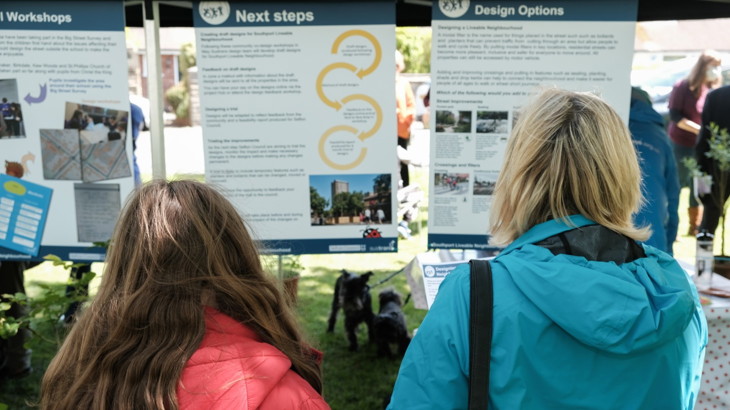 Two women in raincoats looking at a notice board with information about the Southport Liveable Neighbourhood project.