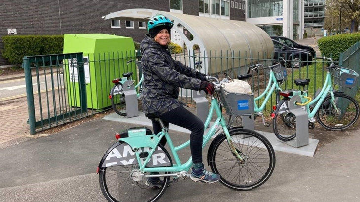 Pregnant woman on a city bike, beside a rack of more city bikes. The bike has a front basket with bags in.