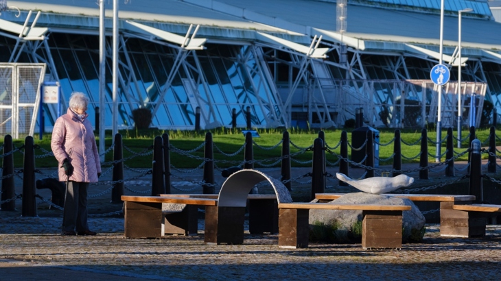 A woman in a winter jacket looks at Allan Potter's Ebb and Flow seating and sculpture at Greenock Waterfront, with the River Clyde in the background. 