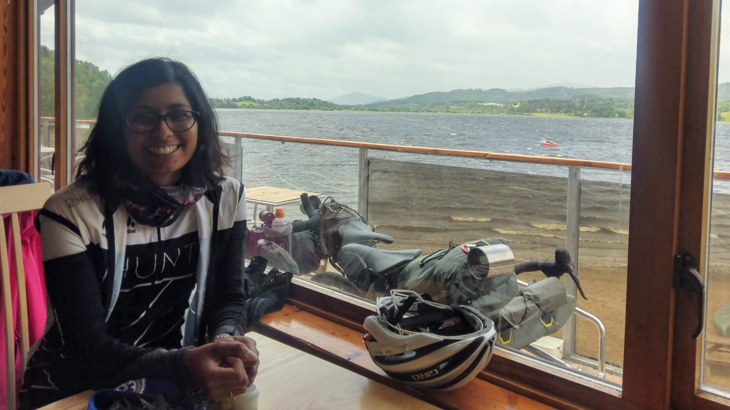 Deepti sat at a cafe table with her bike propped up outside the window, a loch in the distance.