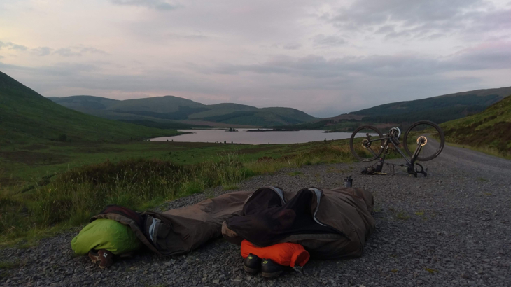 Two bivvy bags set up in a layby beside a loch with mountains all around.