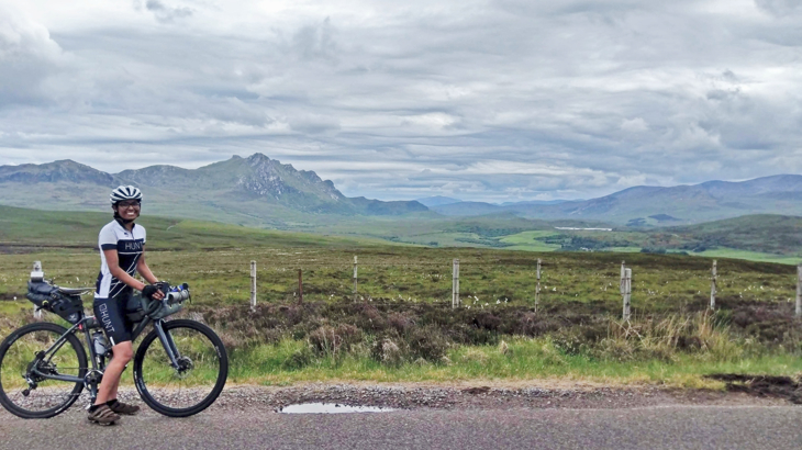Deepti smiling on her bike fitted with bags, on a gravel road beside open countryside, mountains and a loch in the distance.