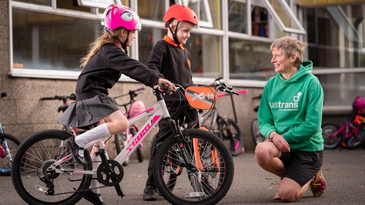 Two children on bikes and wearing cycle helmets, get a lesson from a Walk Wheel Cycle Trust Active School Travel Officer who is kneeling to talk to them.