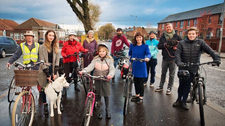 Group of residents stand with bikes and dogs with politician at side of road.