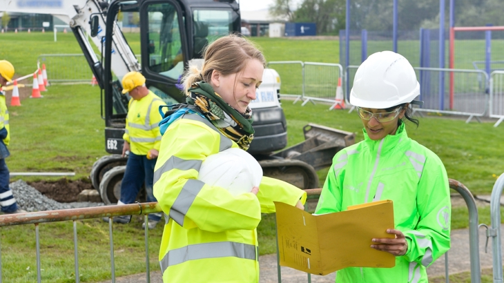 Construction site in a field. Two females in foreground looking at paperwork, wearing hivis and safety wear