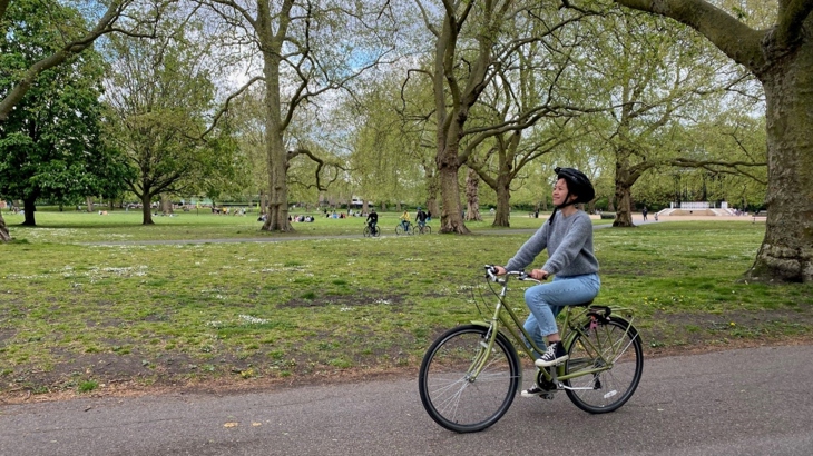 Ying cycles through a leafy tree-lined park, whilst learning to ride a bike.