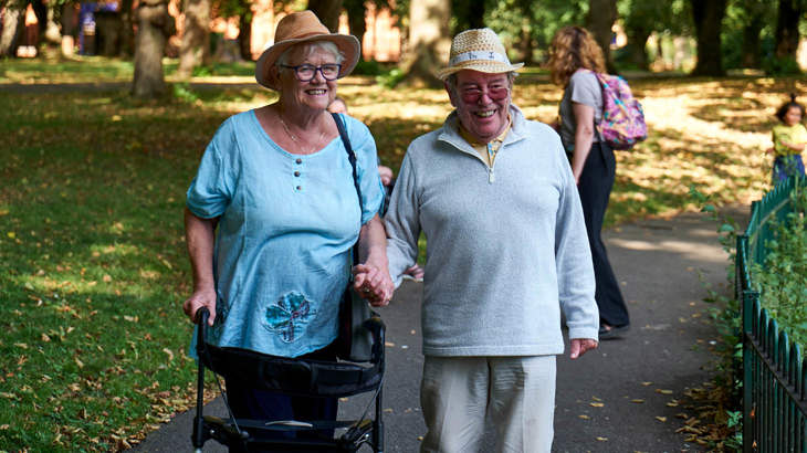 Two older people, one with a frame, smiling as they stroll through a park