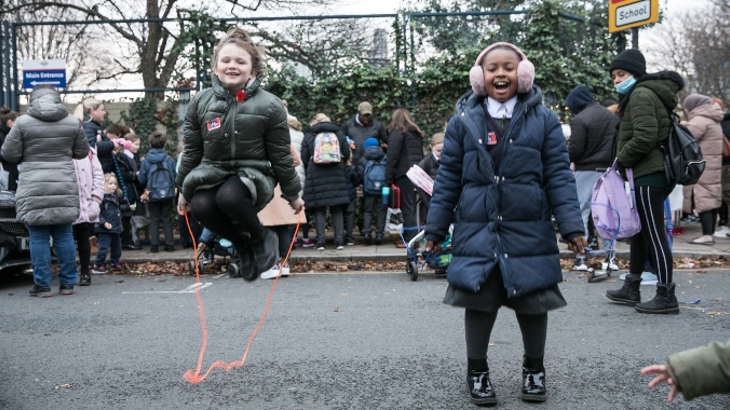 Two laughing primary age children in winter coats skip with ropes outside at a London school.