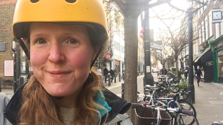 Social Prescribing Link Worker Emma takes a selfie in a London street. She wears a yellow cycling helmet and a row of bikes are parked in the street behind her.