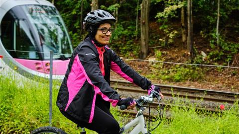 A person wearing a black and pink cycling jacket and helmet rides a bike alongside a tram track, with a pink tram passing through a wooded area.