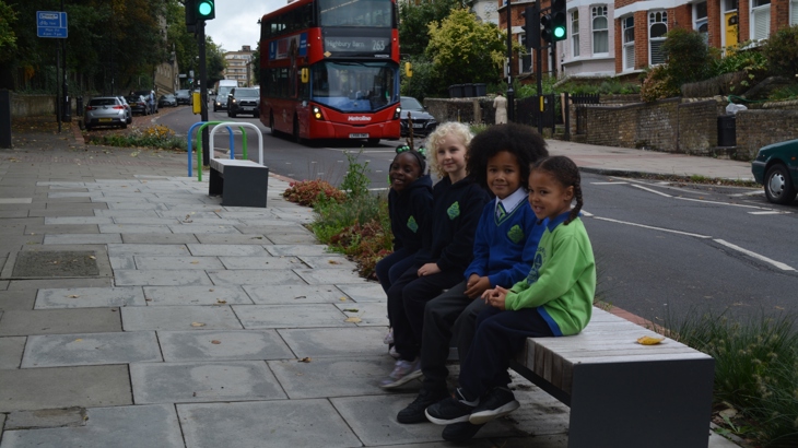 four children sit on a bench beside a busy road