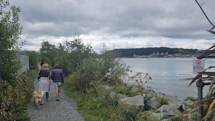 Two adults walking along NCN 5 besides Afon Conwy with a dog and Conwy Castle in the distance.
