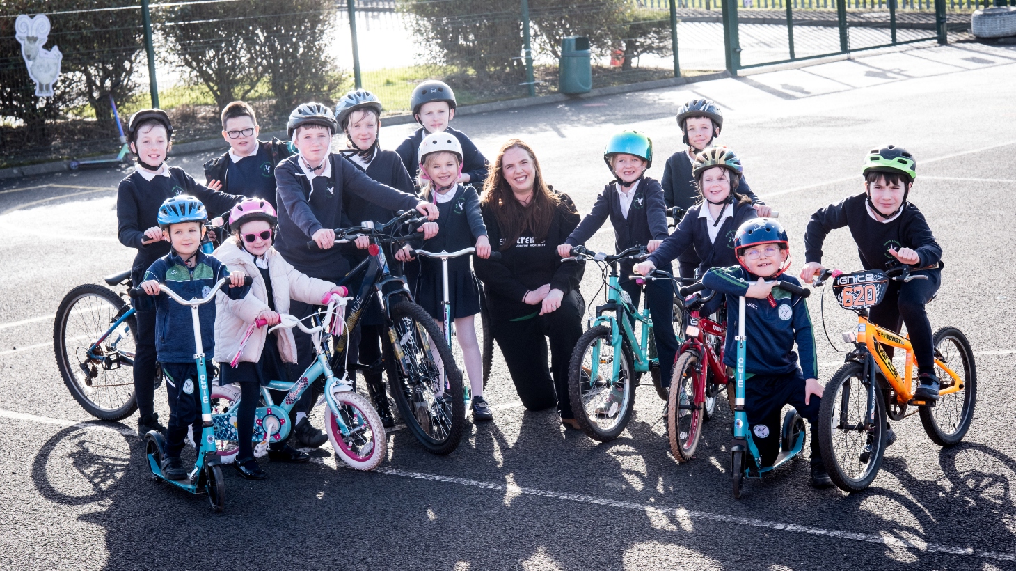 A group of children stand in a school playground with a female Walk Wheel Cycle Trust active travel officer kneeling in the middle of them. Most of the children are on bikes or scooters and are wearing helmets.