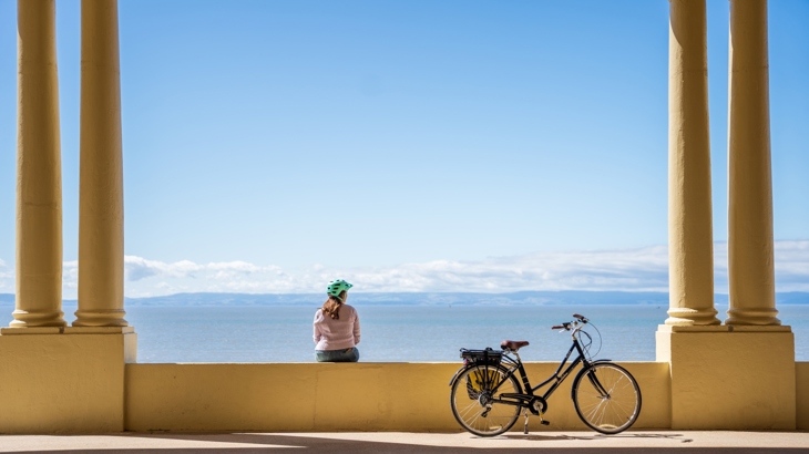 A woman sits on Barry Island promenade looking away from the camera toward the sea. Beside her is a parked e-bike.