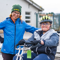 A woman stood next to a young boy sat on a trike wearing a helmet they are both looking at the camera and smiling 