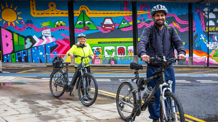 Two people stood smiling, wearing helmets with their bikes in front of a bright art mural in Scotland