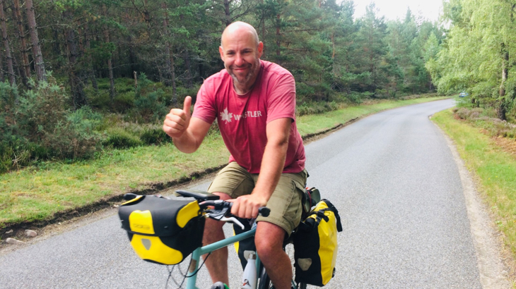 A white man with a bald head riding his bike through a road lined with thick trees while smiling and giving a thumbs up 